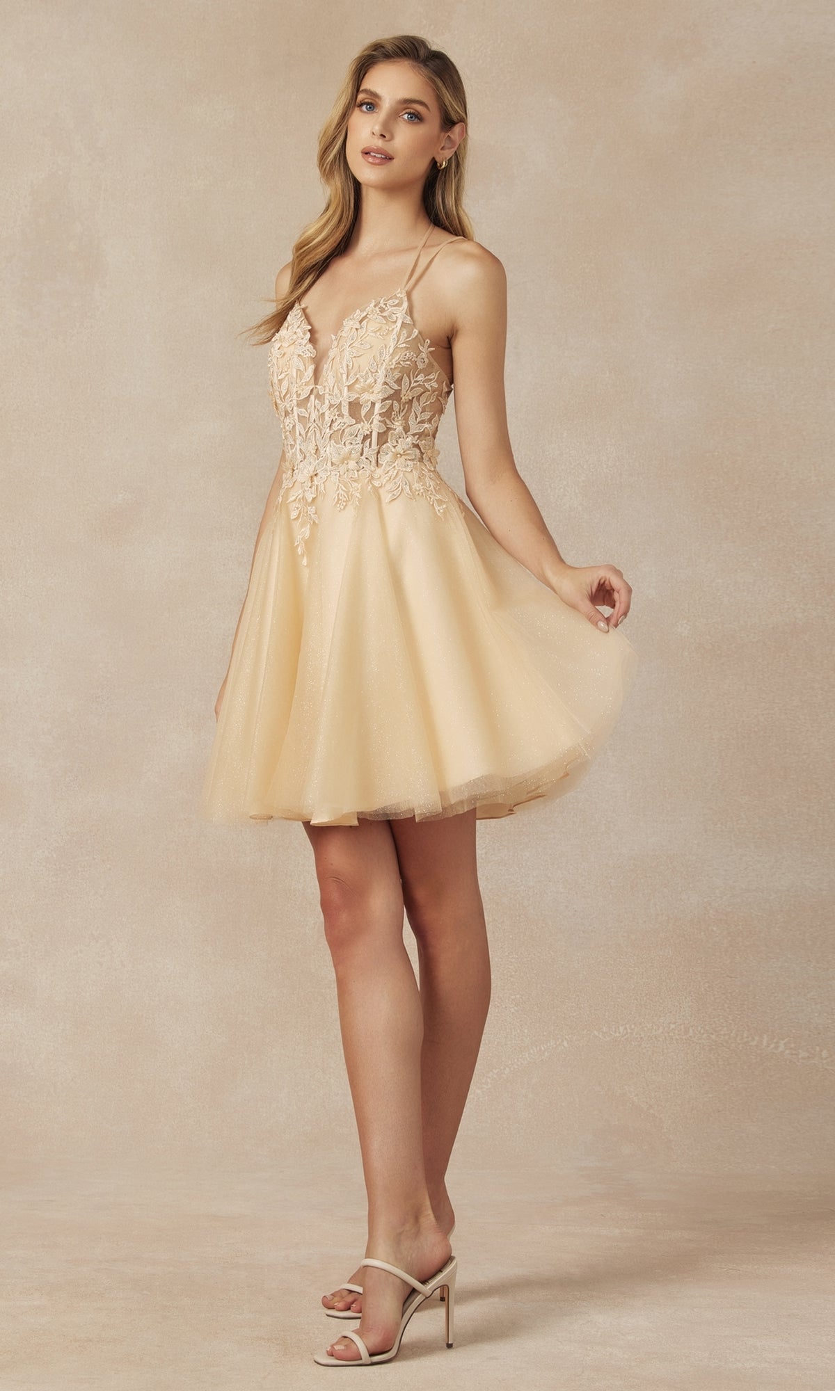 A woman wears the Glitter-Tulle Short A-Line Homecoming Dress 864, featuring spaghetti straps, a lace bodice, and a flared tulle skirt in light beige, standing before a neutral background.